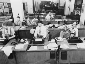 Black and white image of newsroom with journalists at desks.