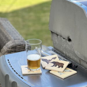 Wooden coasters with a glass of beer on a barbecue with images of salmon, bear, bat and eagle.